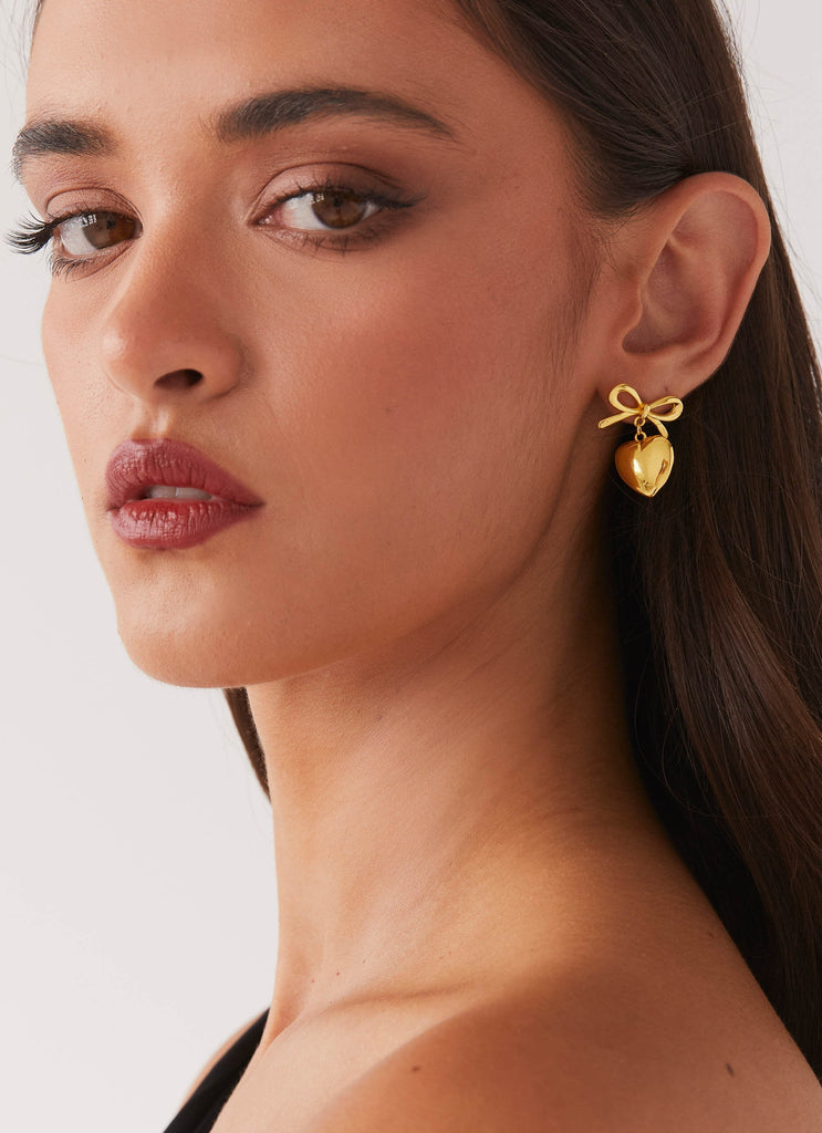 Womens Chasity Bow And Heart Earrings in the colour Gold in front of a light grey background